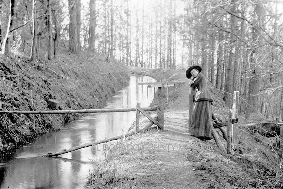 Historic black-and-white photograph of a woman beside the El Dorado Canal, with wooden railings and a forested hillside.
