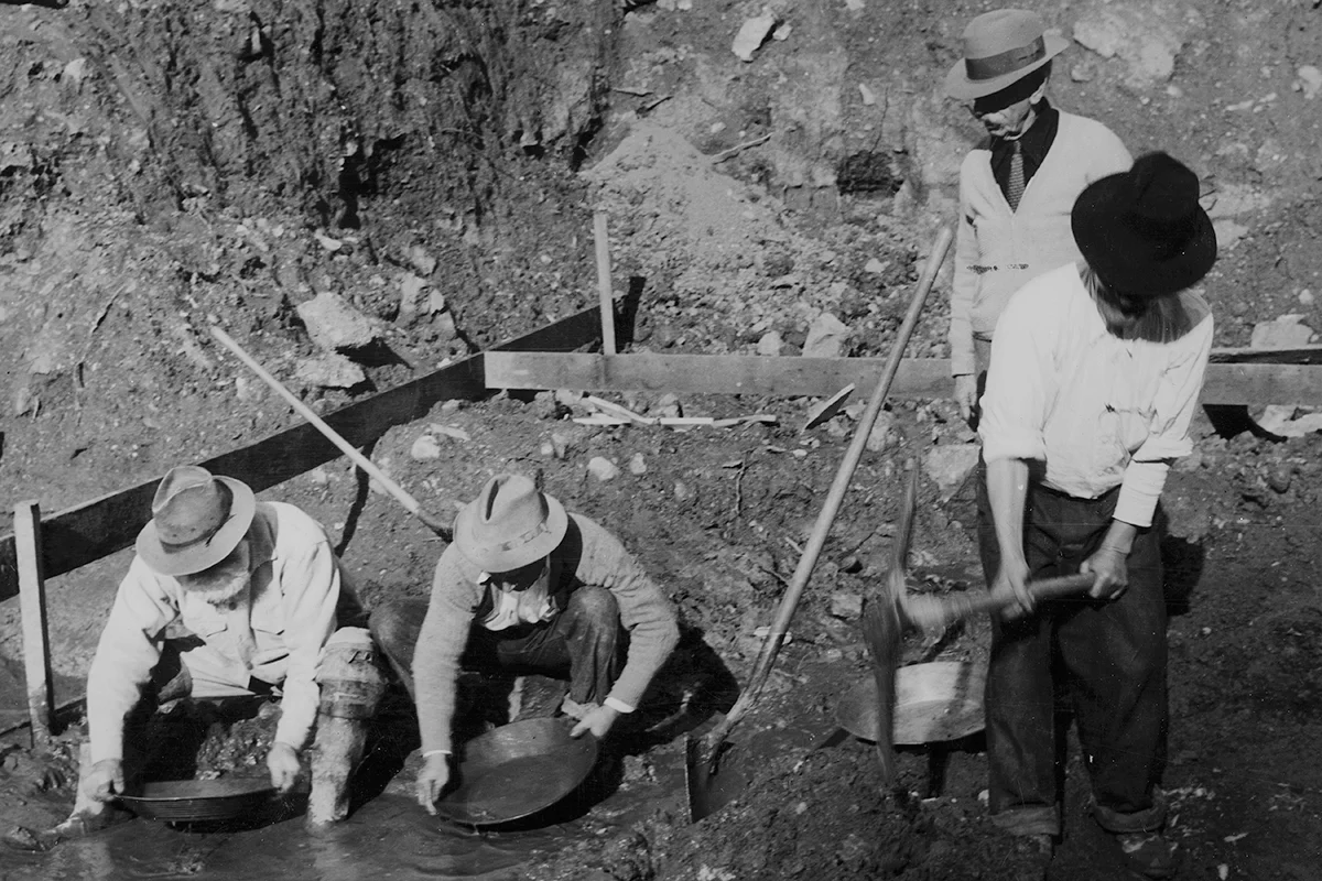 Black-and-white photograph of two men panning for gold while another works with a pickaxe and a fourth looks on.