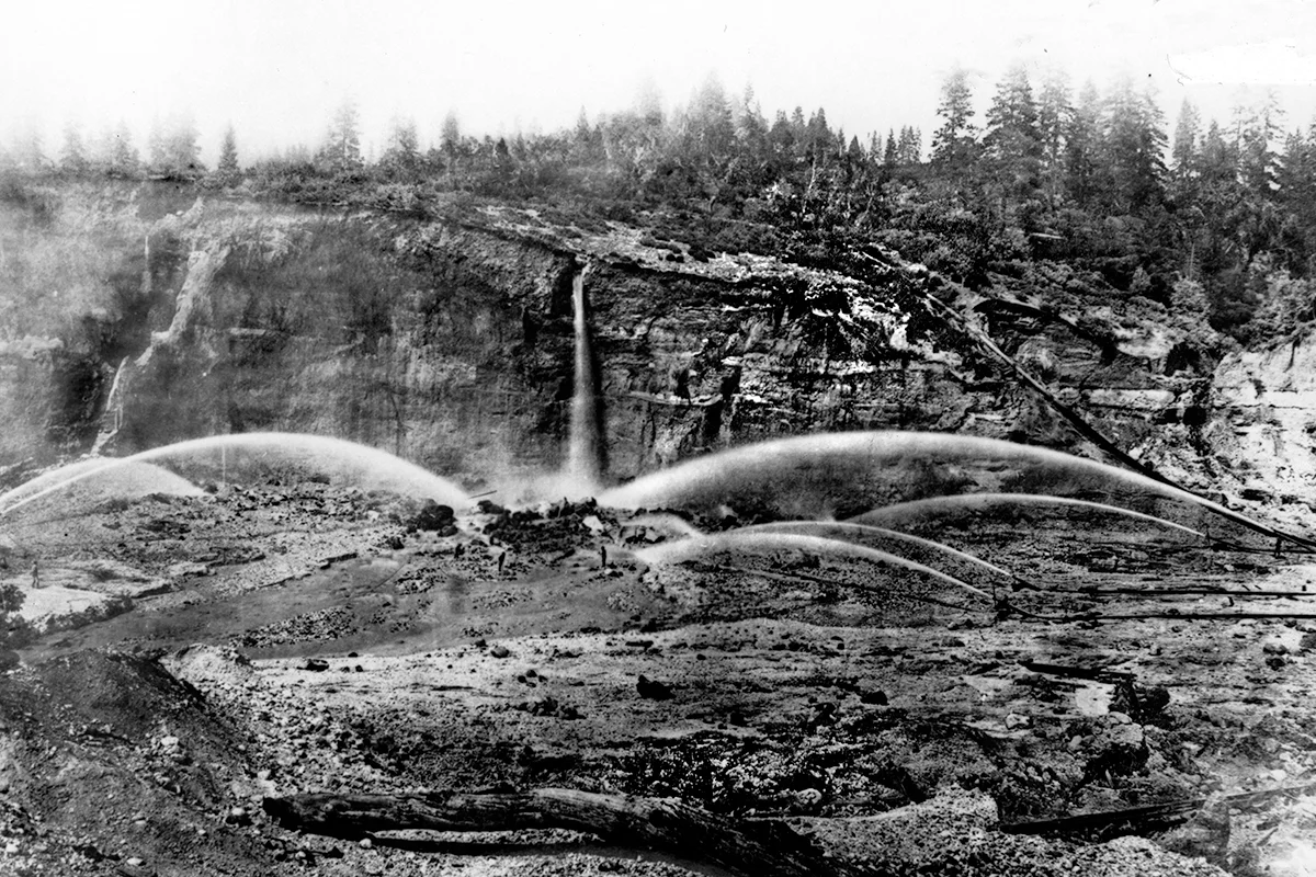 Black-and-white photograph of hydraulic mining at Big Cut, with water jets spraying exposed earth below tree-lined cliffs.